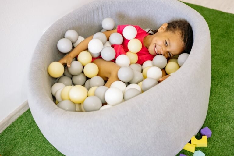 Happy child playing in a soft ball pit filled with gray, white, and yellow balls at an indoor play area.