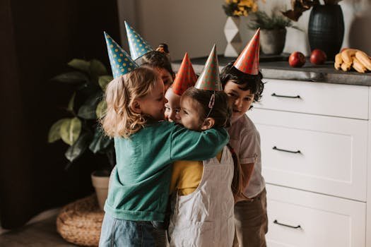 Four kids wearing party hats and hugging warmly during a birthday celebration indoors.